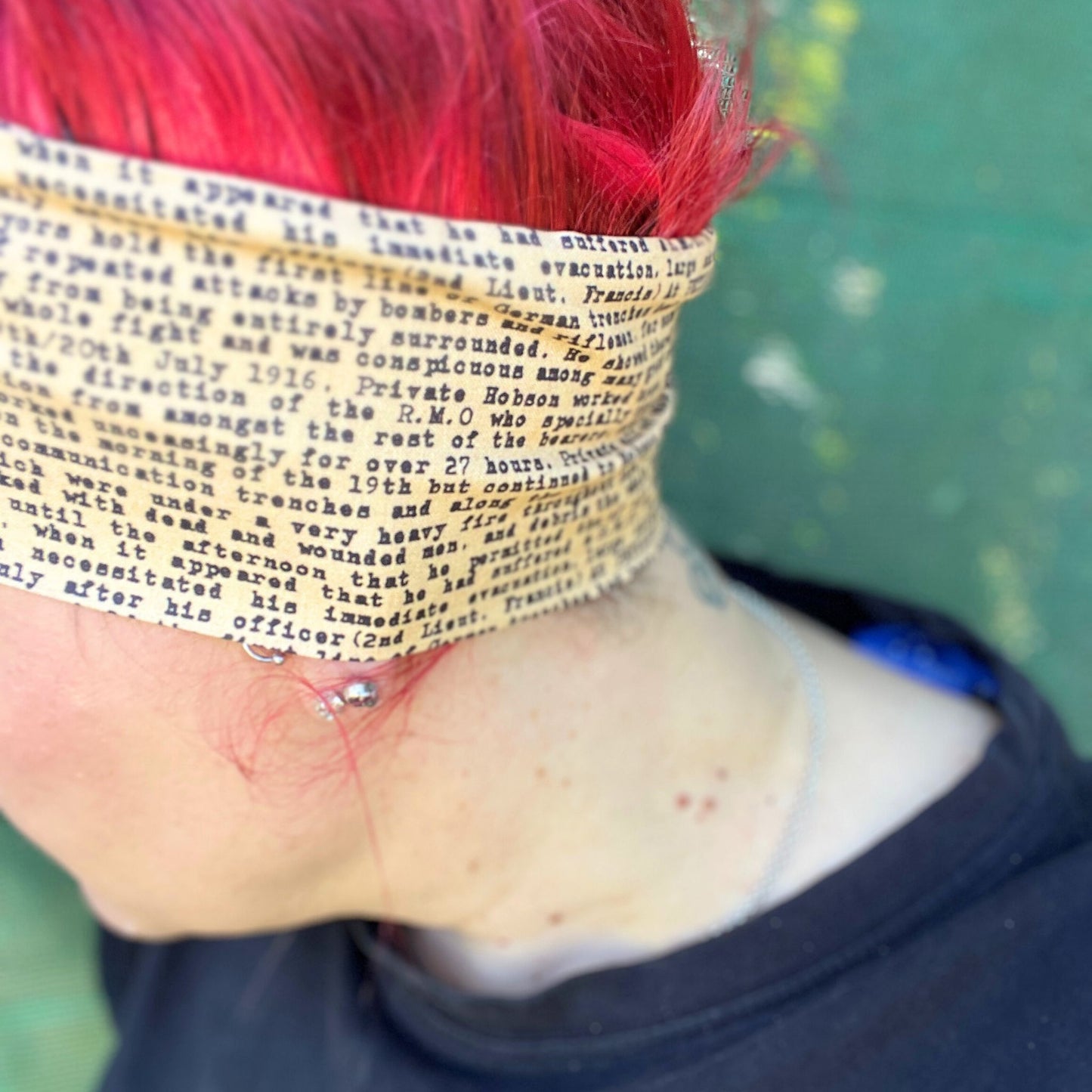 In this image, a woman sports a vibrant red hairstyle, complementing it with a wire headwrap that features an ANZAC soldier's letter a unique print. Adding a historical touch to the headwrap’s design. The headwrap is tied at the top, giving it a bow-like appearance, which accentuates its stylish and laid-back vibe. The woman is looking down, showcasing the headwrap as the standout accessory.