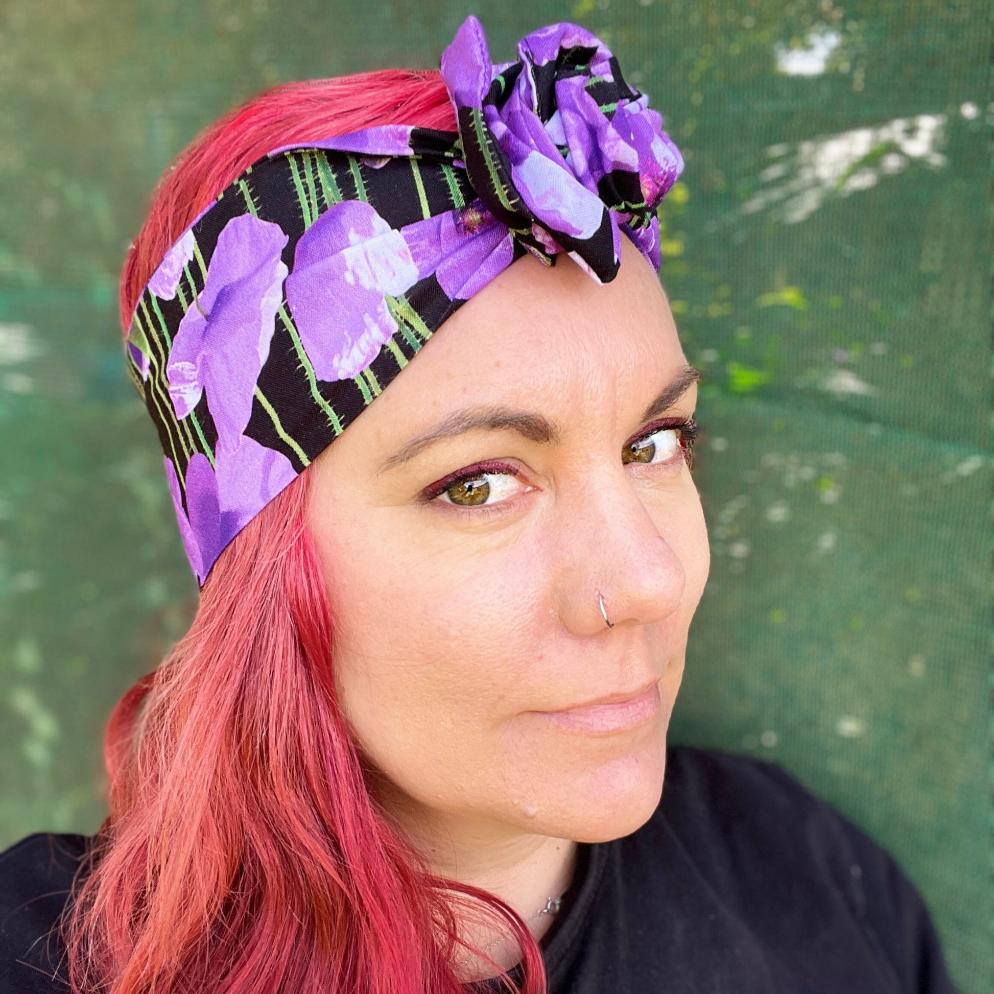 showcasing a wire headwrap adorned with bold purple poppies against a black background. The headwrap is tied in a charming knot atop her head, lending a playful and stylish look. The vivid purple of the flowers stands out