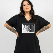 A woman with long dark hair and visible arm tattoos wears an oversized black V-neck T-shirt with a bold white geometric logo on the chest, posed against a light gray studio background.
