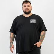 A man with a beard and tattooed arms wearing a black v-neck t-shirt with a white graphic logo on the left chest, standing against a light gray background.