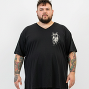 A man with a beard and tattooed arms wearing a black v-neck t-shirt with a white raccoon graphic on the left chest, photographed against a light gray background.