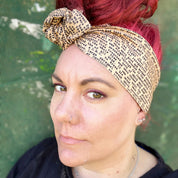 In this image, a woman sports a vibrant red hairstyle, complementing it with a wire headwrap that features an ANZAC soldier's letter a unique print. Adding a historical touch to the headwrap’s design. The headwrap is tied at the top, giving it a bow-like appearance, which accentuates its stylish and laid-back vibe. The woman is looking down, showcasing the headwrap as the standout accessory.