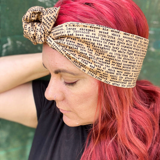 In this image, a woman sports a vibrant red hairstyle, complementing it with a wire headwrap that features an ANZAC soldier's letter a unique print. Adding a historical touch to the headwrap’s design. The headwrap is tied at the top, giving it a bow-like appearance, which accentuates its stylish and laid-back vibe. The woman is looking down, showcasing the headwrap as the standout accessory.