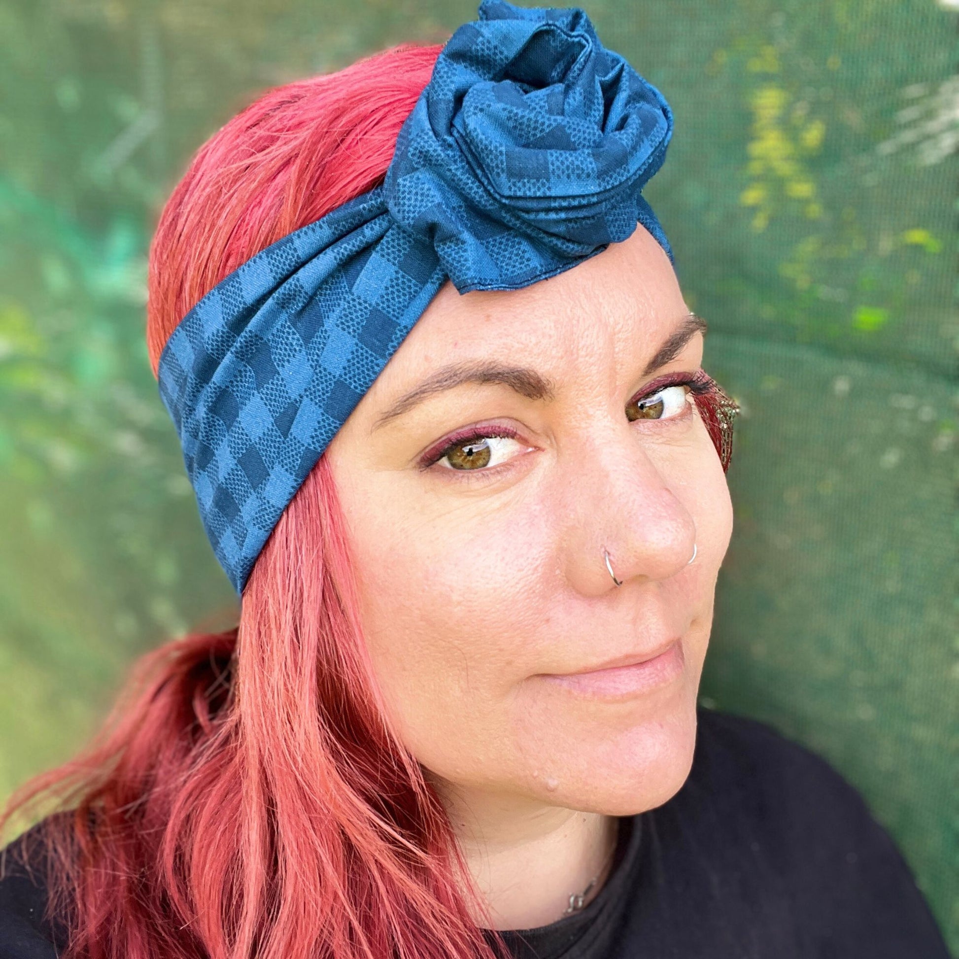 showcasing a stylish blue check-patterned wire headwrap. The headwrap features a knotted bow on the top, adding a playful twist to the design. Her vibrant red hair peeks out beneath the headwrap, creating a striking contrast against the blue. 