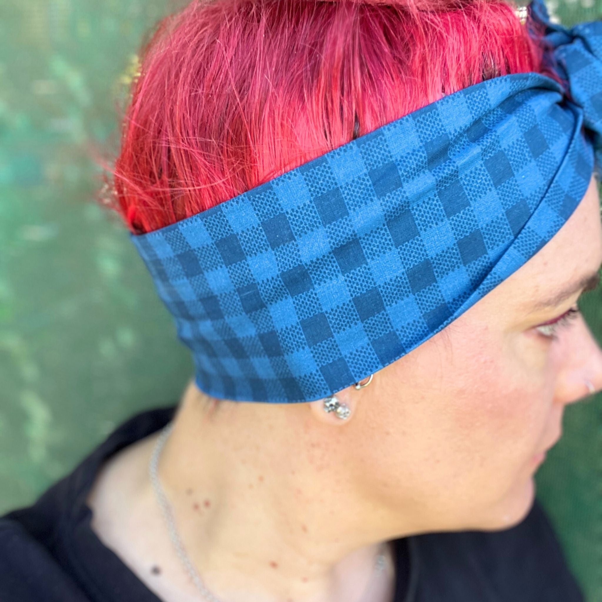 showcasing a stylish blue check-patterned wire headwrap. The headwrap features a knotted bow on the top, adding a playful twist to the design. Her vibrant red hair peeks out beneath the headwrap, creating a striking contrast against the blue. 