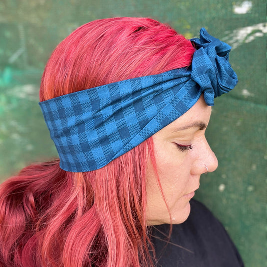 showcasing a stylish blue check-patterned wire headwrap. The headwrap features a knotted bow on the top, adding a playful twist to the design. Her vibrant red hair peeks out beneath the headwrap, creating a striking contrast against the blue. 