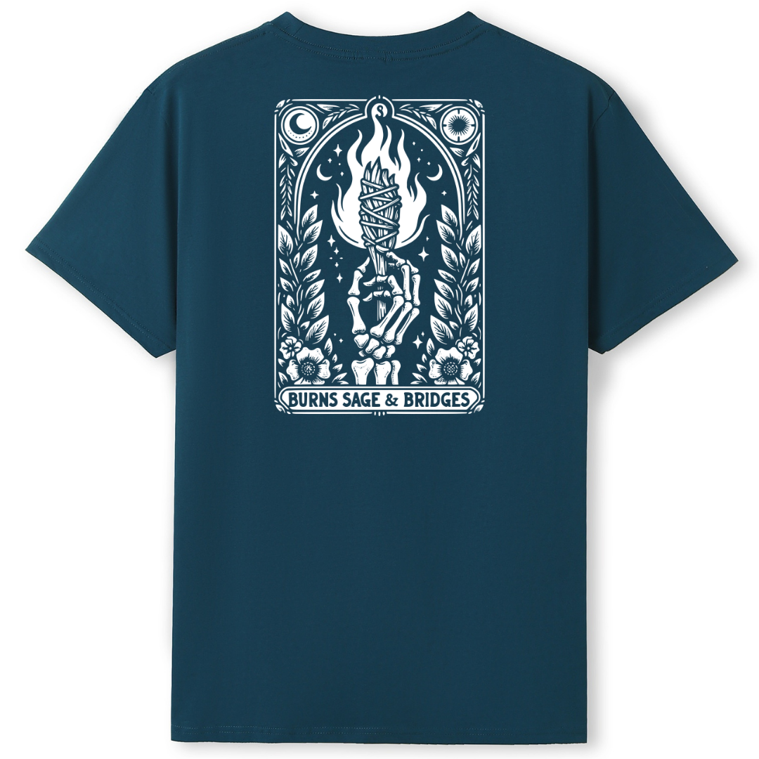 A dark teal blue short-sleeve t-shirt with a large white graphic print on the back. The graphic features a tarot card design with a hand holding a flaming torch, surrounded by a laurel wreath, stars, and moons, with the text 'BURNS SAGE & BRIDGES' at the bottom.