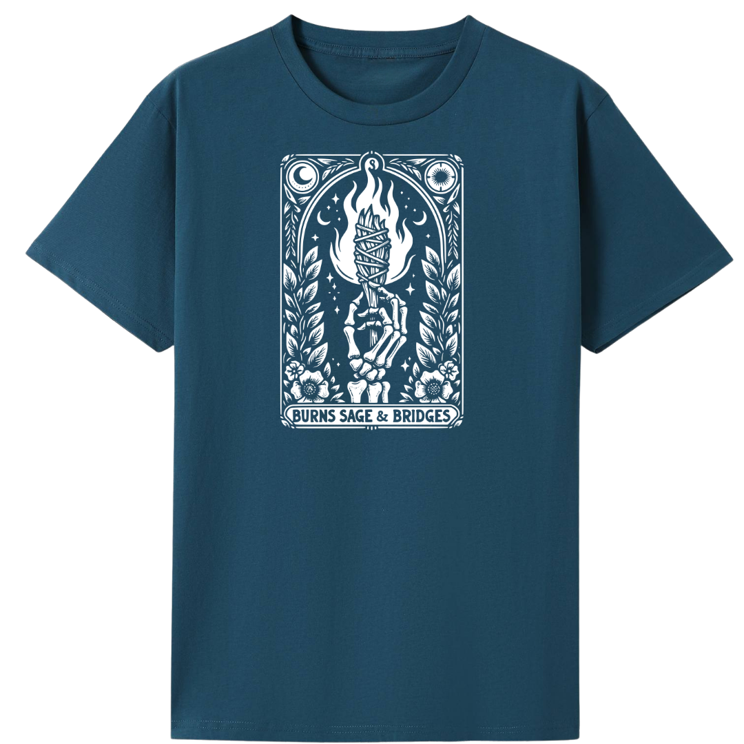 A dark teal blue short-sleeve t-shirt with a white graphic print on the chest. The graphic features a stylized illustration of a hand holding a lit candle with flames, surrounded by leaves and celestial symbols, with the text 'BURNS SAGE & BRIDGES' at the bottom.