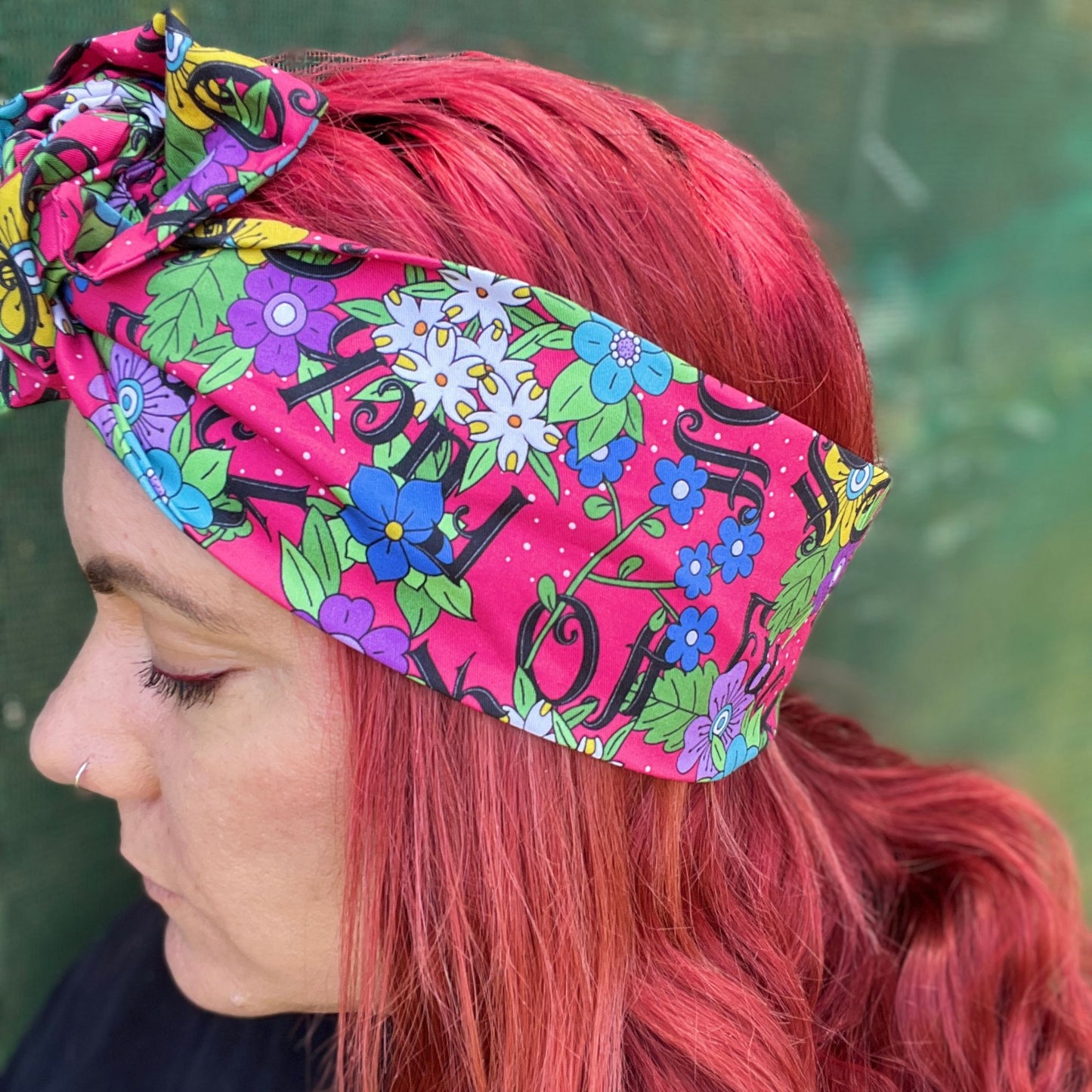 The headwrap boasts a lively floral pattern with a mix of bright flowers and leaves on a hot pink background, interspersed with bold, playful text. It's knotted at the top, giving a touch of retro flair. 