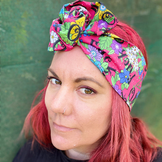 The headwrap boasts a lively floral pattern with a mix of bright flowers and leaves on a hot pink background, interspersed with bold, playful text. It's knotted at the top, giving a touch of retro flair. 
