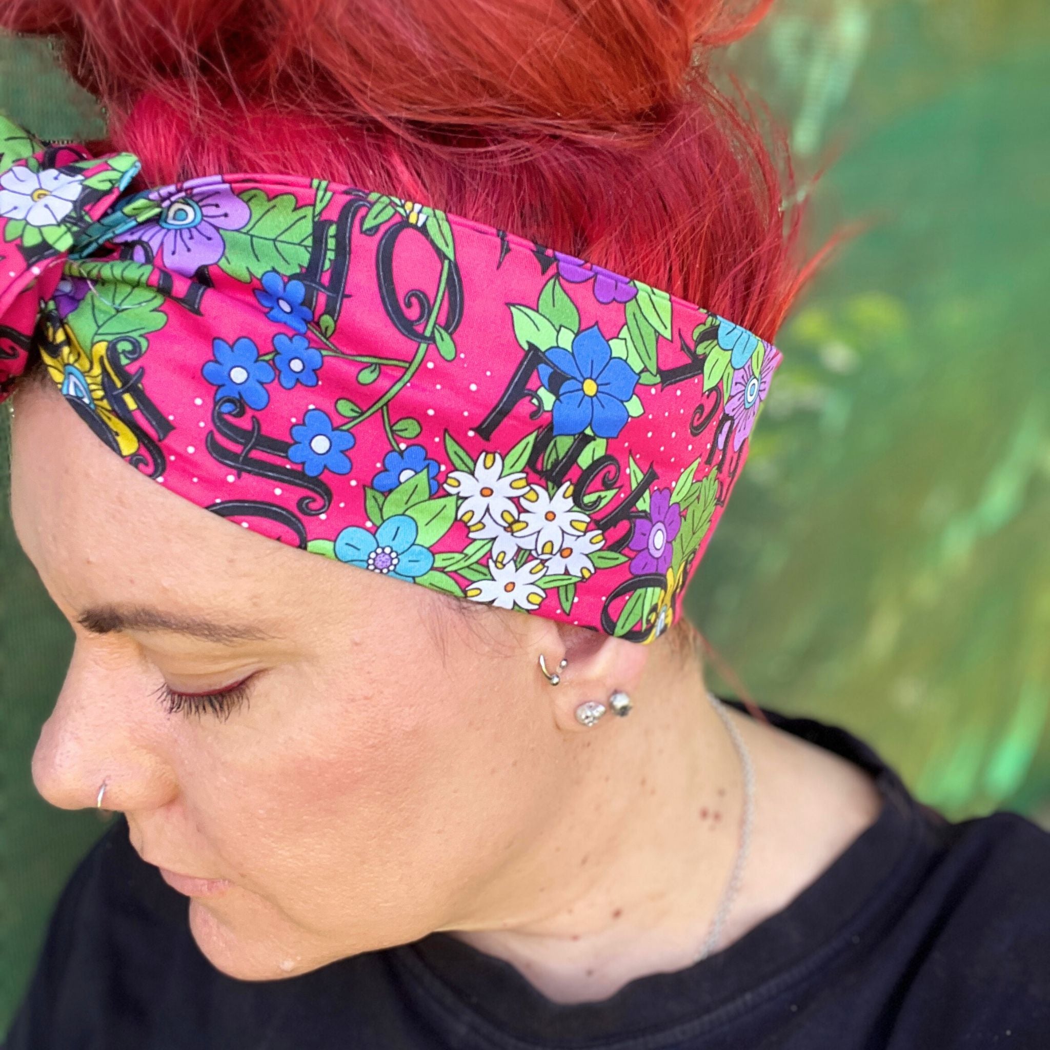 The headwrap boasts a lively floral pattern with a mix of bright flowers and leaves on a hot pink background, interspersed with bold, playful text. It's knotted at the top, giving a touch of retro flair. 
