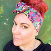 The headwrap boasts a lively floral pattern with a mix of bright flowers and leaves on a hot pink background, interspersed with bold, playful text. It's knotted at the top, giving a touch of retro flair. 