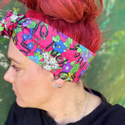 The headwrap boasts a lively floral pattern with a mix of bright flowers and leaves on a hot pink background, interspersed with bold, playful text. It's knotted at the top, giving a touch of retro flair. 