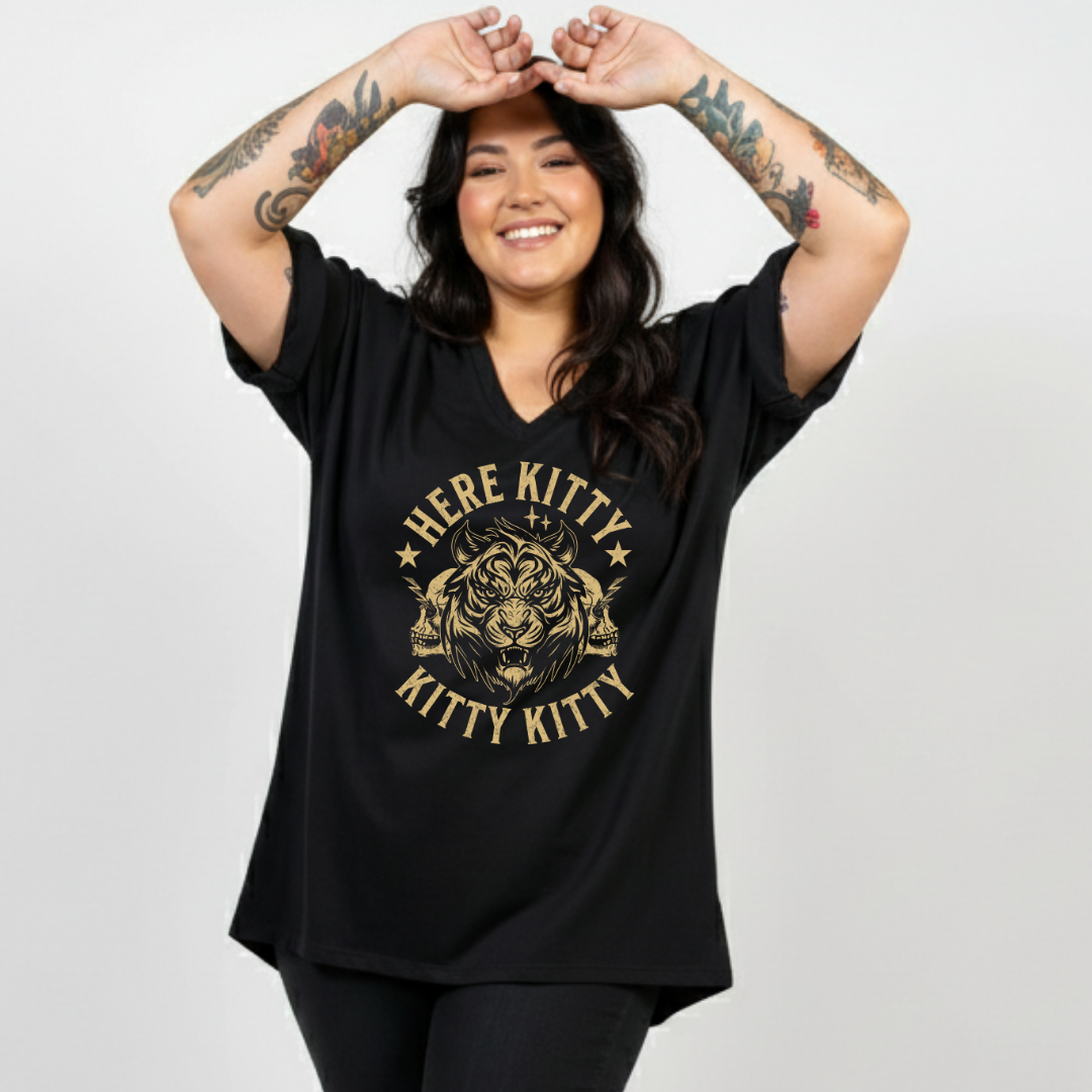 A woman with tattooed arms wearing a black oversized V-neck t-shirt with a gold graphic of a cat head and the text 'HERE KITTY KITTY KITTY'.