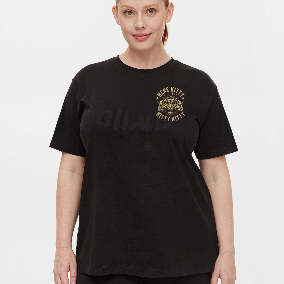 A woman modeling a black short-sleeve t-shirt with a gold graphic on the left chest that reads 'HERE KITTY KITTY KITTY' around a kitten illustration. The word 'SHAPES' is printed in a subtle, tonal black font across the center chest.