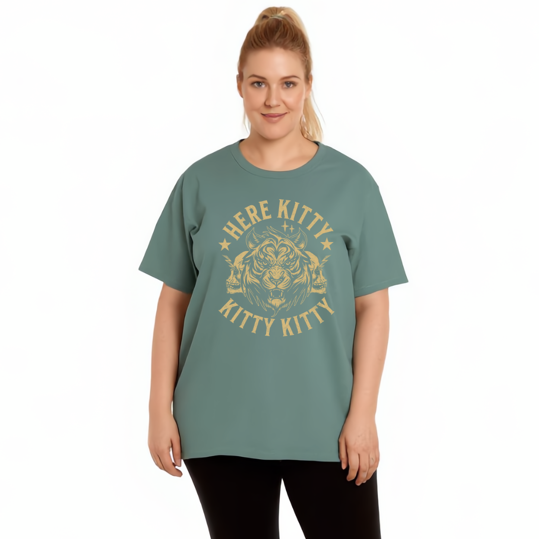A woman modeling a sage green short-sleeve t-shirt with a distressed gold graphic that reads 'HERE KITTY KITTY' around a cat face illustration, set against a plain white background.