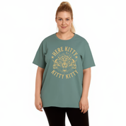 A woman modeling a sage green short-sleeve t-shirt with a distressed gold graphic that reads 'HERE KITTY KITTY' around a cat face illustration, set against a plain white background.