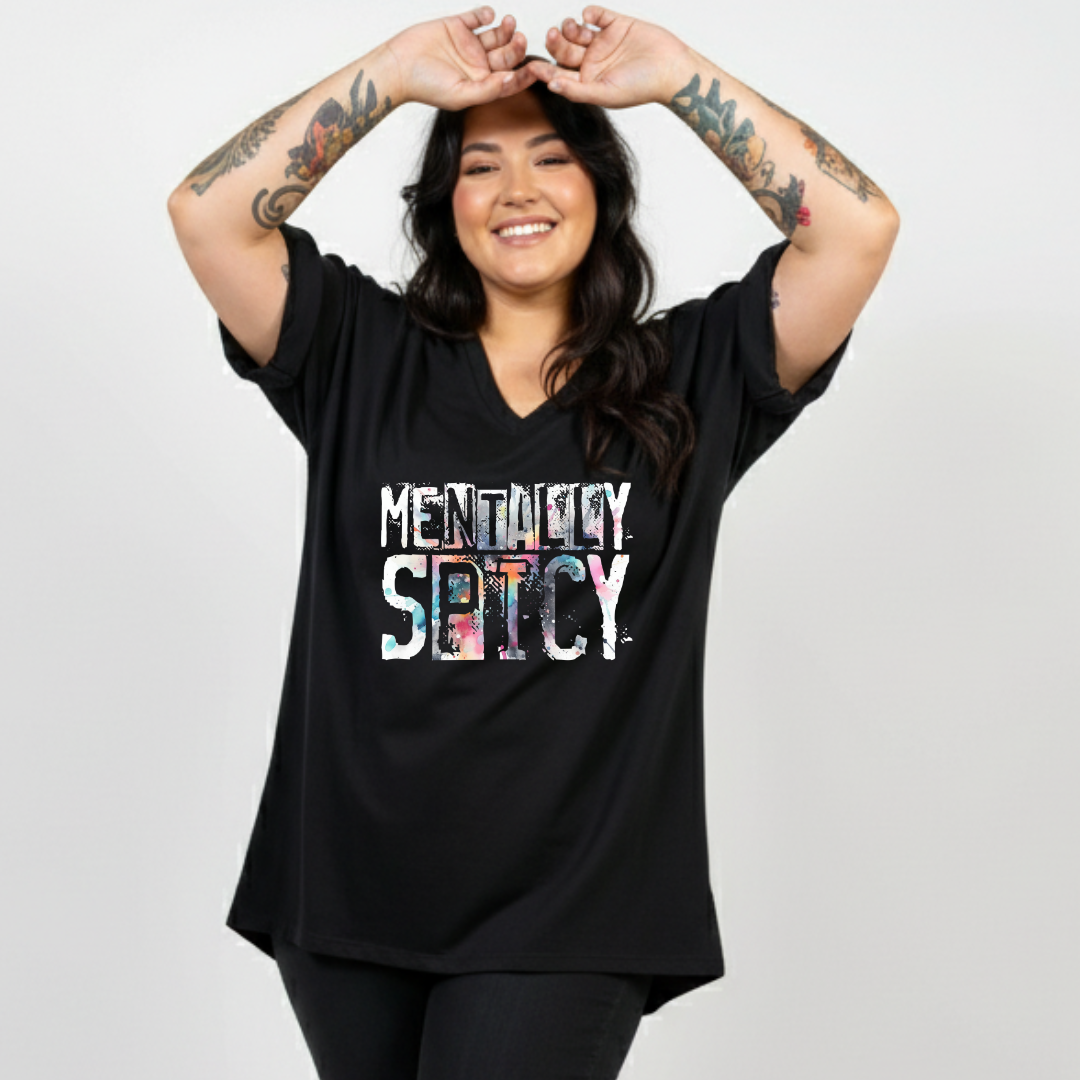 A woman with tattoos on her arms is wearing a black V-neck t-shirt with a colorful graphic that reads 'MENTALITY SPICY'. She is posing with her hands above her head against a light gray studio background.