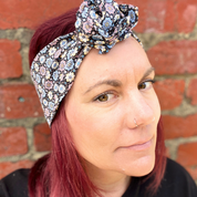 Woman with red hair wearing a floral headscarf against a brick wall.