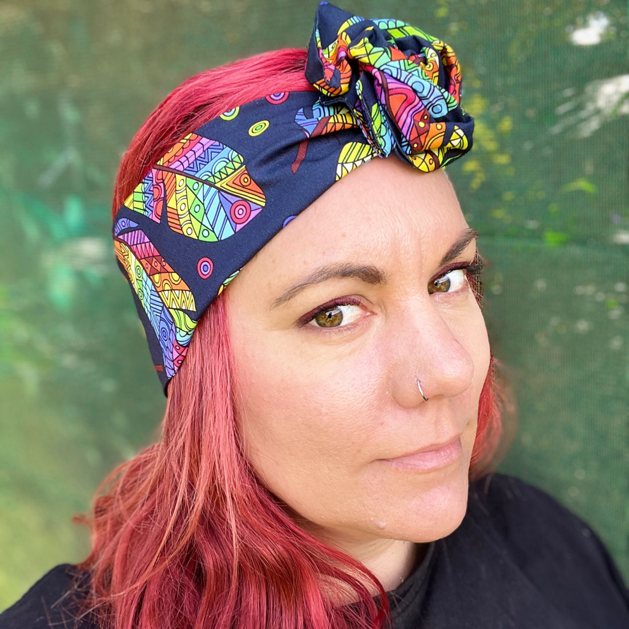 wearing a headwrap with a striking Maori-inspired feather design. The pattern is vibrant, showcasing a spectrum of colors like yellow, red, green, and blue on a dark background