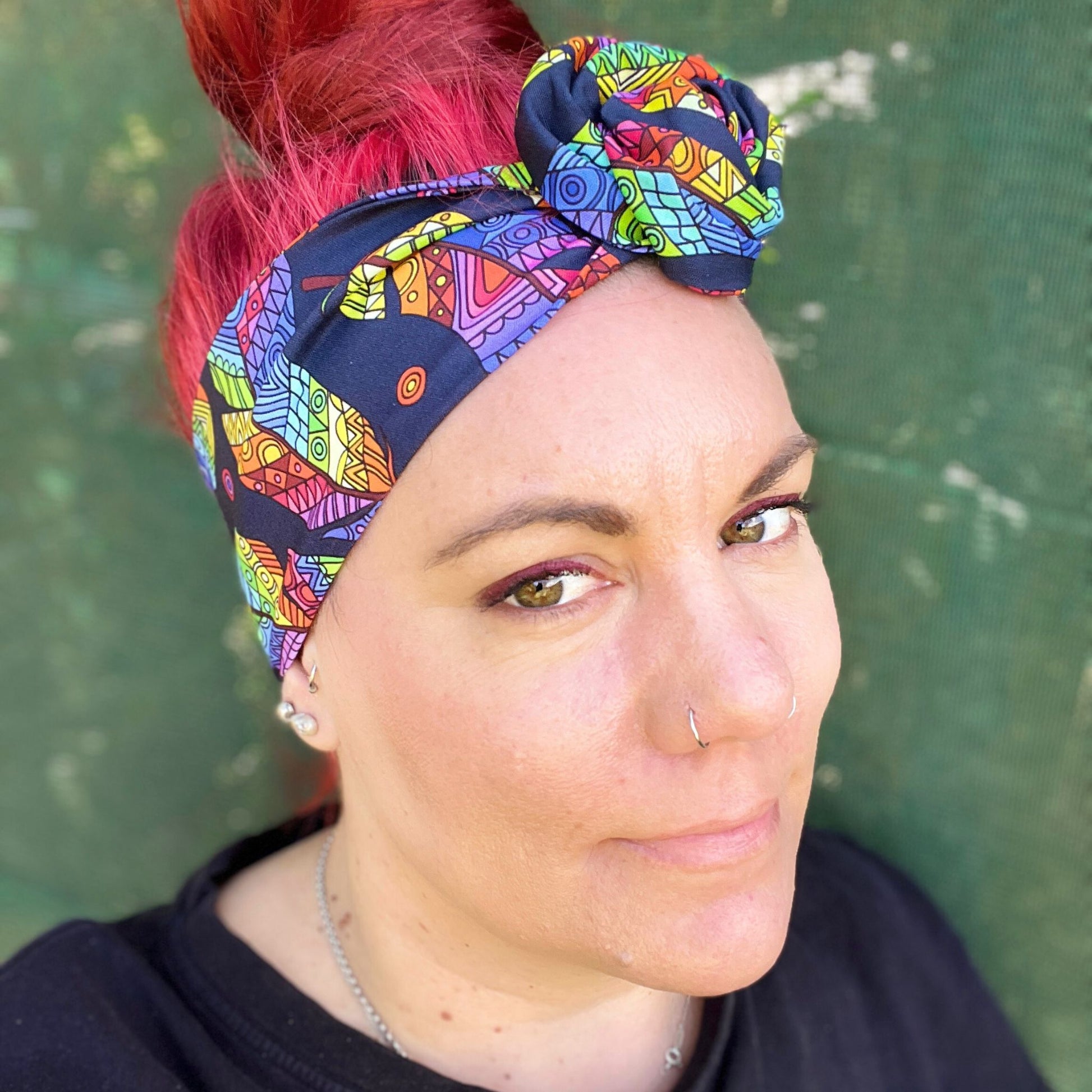 wearing a headwrap with a striking Maori-inspired feather design. The pattern is vibrant, showcasing a spectrum of colors like yellow, red, green, and blue on a dark background