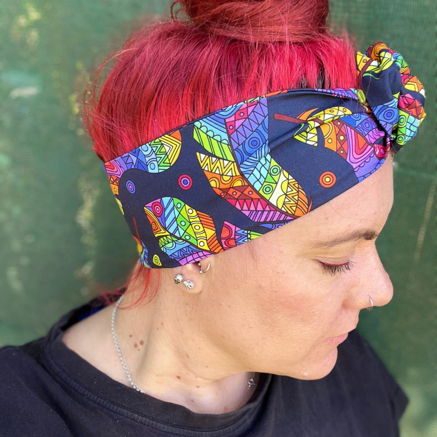 wearing a headwrap with a striking Maori-inspired feather design. The pattern is vibrant, showcasing a spectrum of colors like yellow, red, green, and blue on a dark background