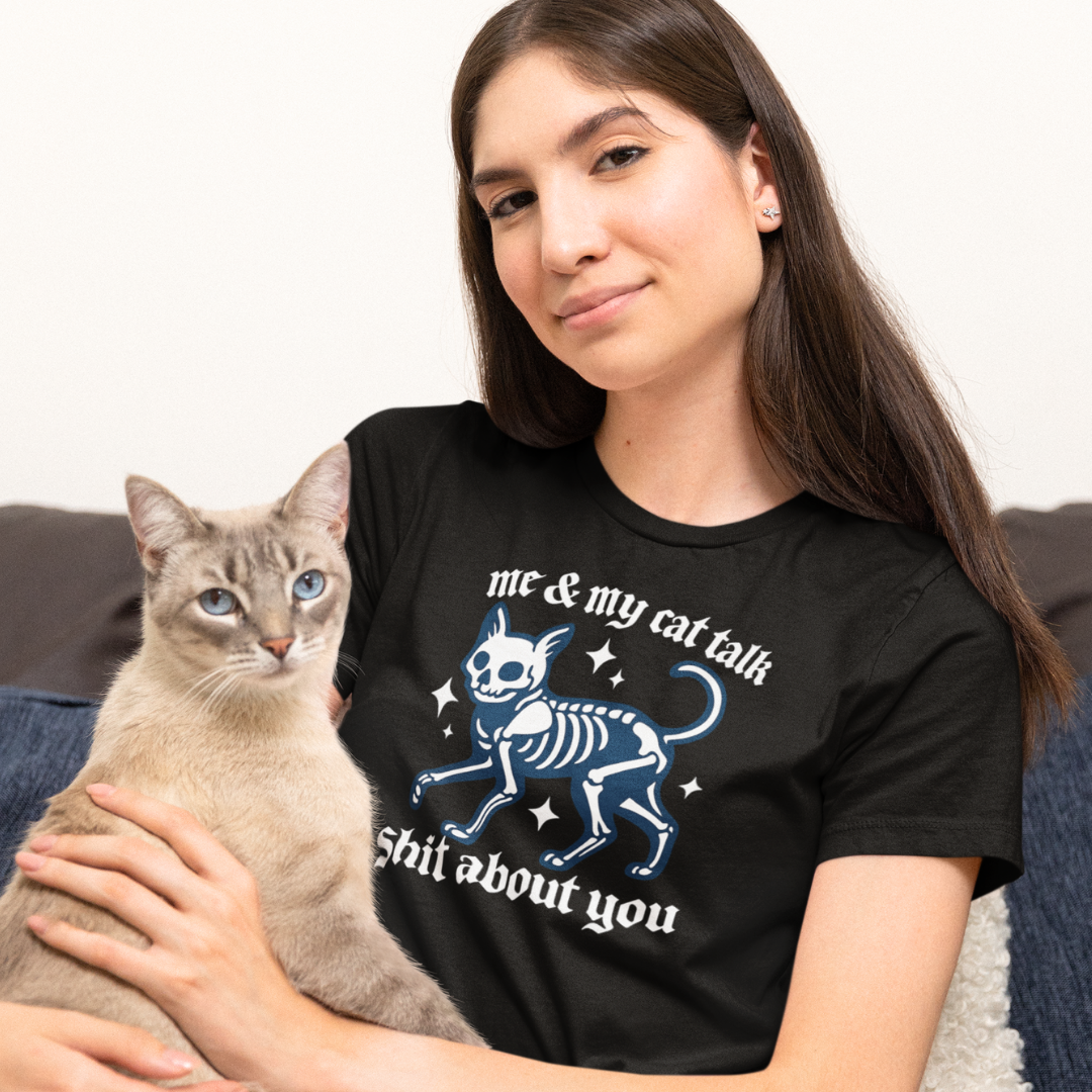 Woman wearing a black t-shirt with a cat skeleton design and text, sitting on a couch with a cat.