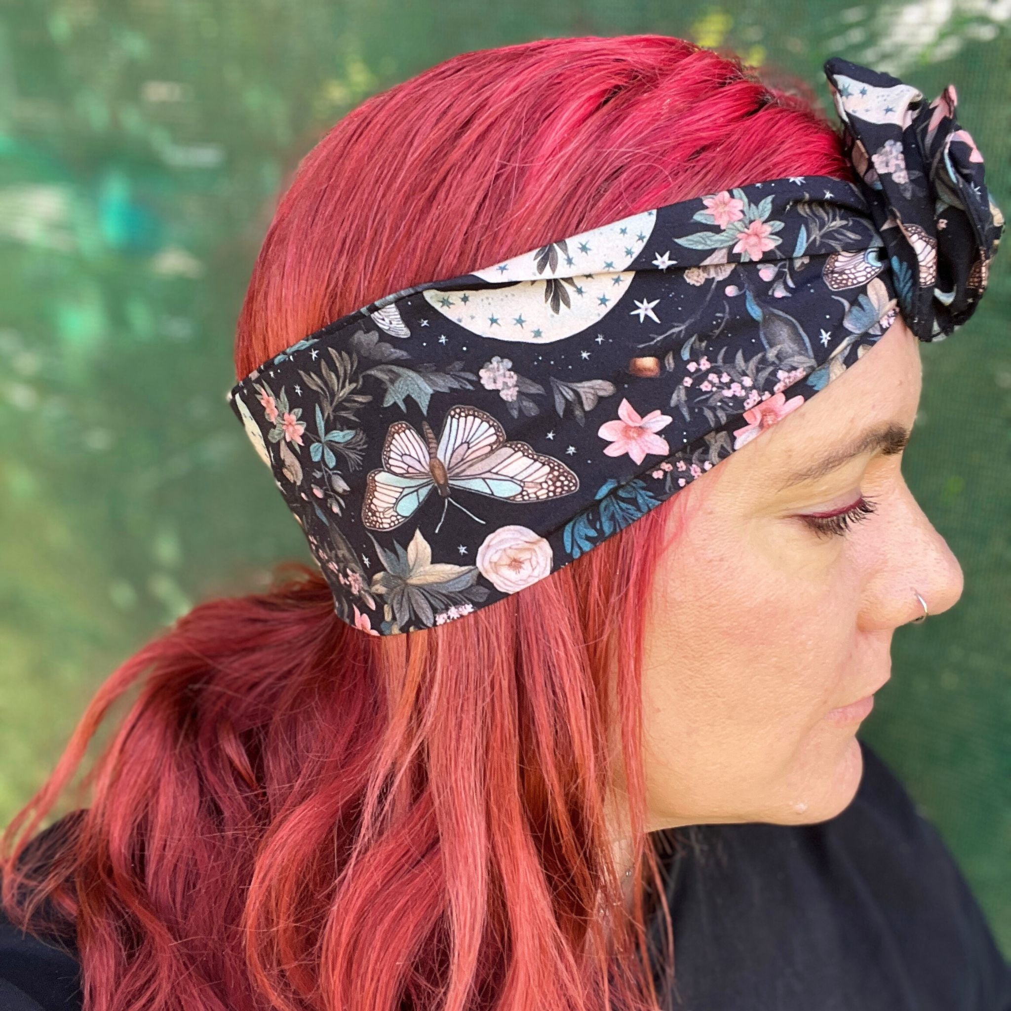  a wire headwrap featuring a mystical garden theme. The headwrap is black and illustrated with delicate butterflies, stars, and an array of soft-colored flowers, creating an enchanting look. It is tied at the top, adding an elegant twist to the design
