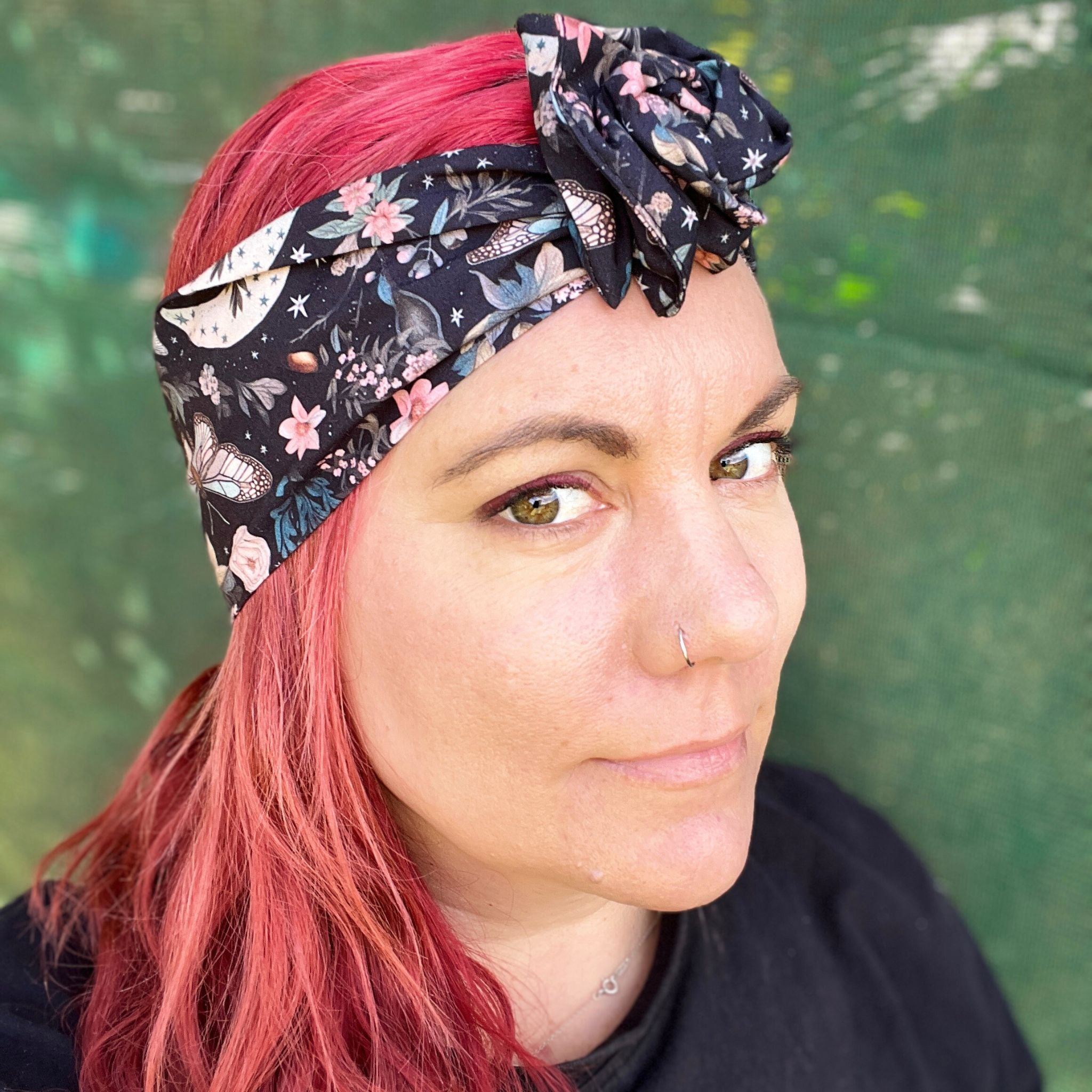  a wire headwrap featuring a mystical garden theme. The headwrap is black and illustrated with delicate butterflies, stars, and an array of soft-colored flowers, creating an enchanting look. It is tied at the top, adding an elegant twist to the design
