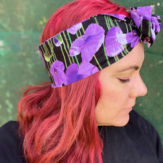 showcasing a wire headwrap adorned with bold purple poppies against a black background. The headwrap is tied in a charming knot atop her head, lending a playful and stylish look. The vivid purple of the flowers stands out