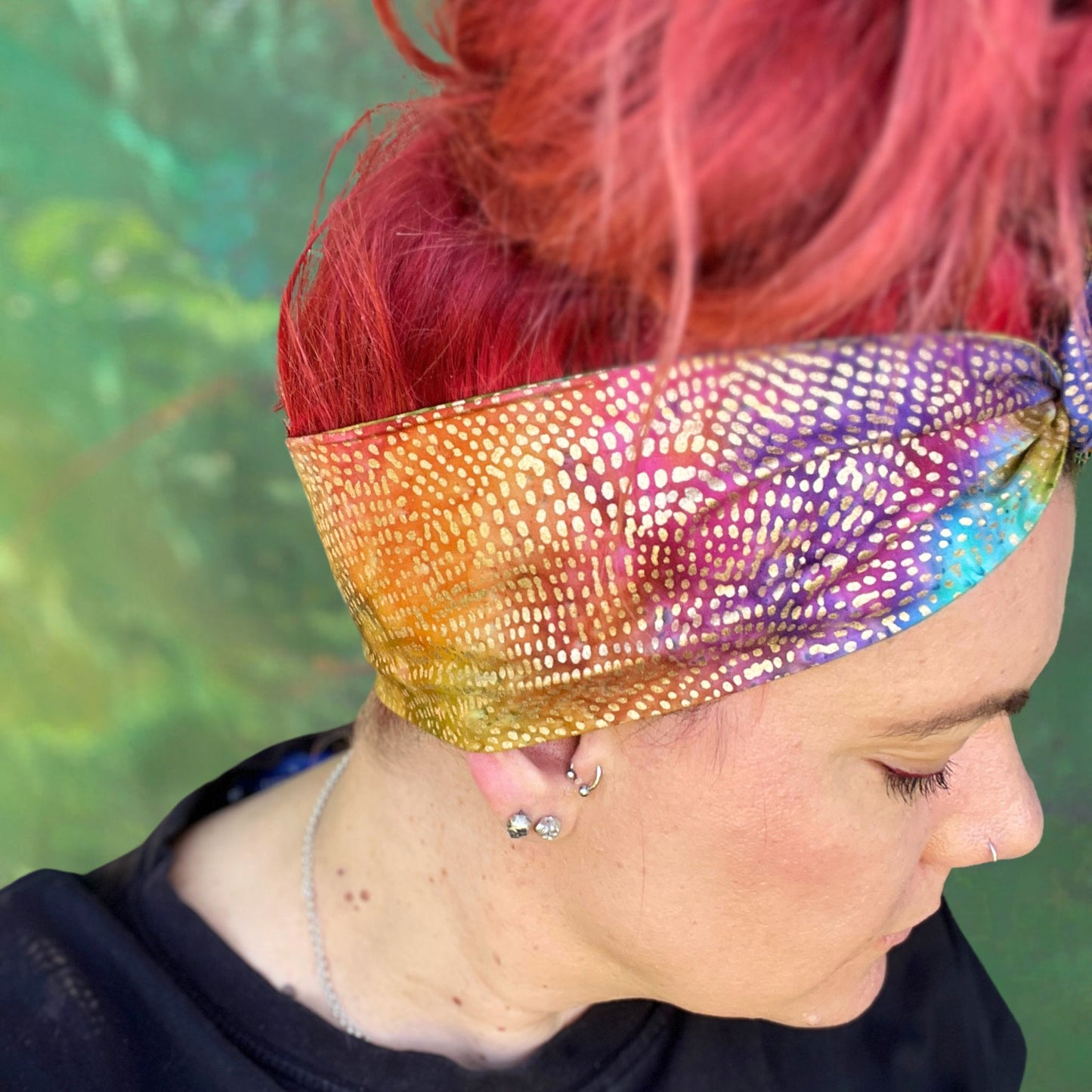 wire headwrap featuring a radiant metallic sheen with a gradient of colors, resembling a rainbow effect. The headwrap is artfully tied in a knot at the side of her head, lending a touch of whimsy to the overall look. 