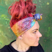 wire headwrap featuring a radiant metallic sheen with a gradient of colors, resembling a rainbow effect. The headwrap is artfully tied in a knot at the side of her head, lending a touch of whimsy to the overall look. 