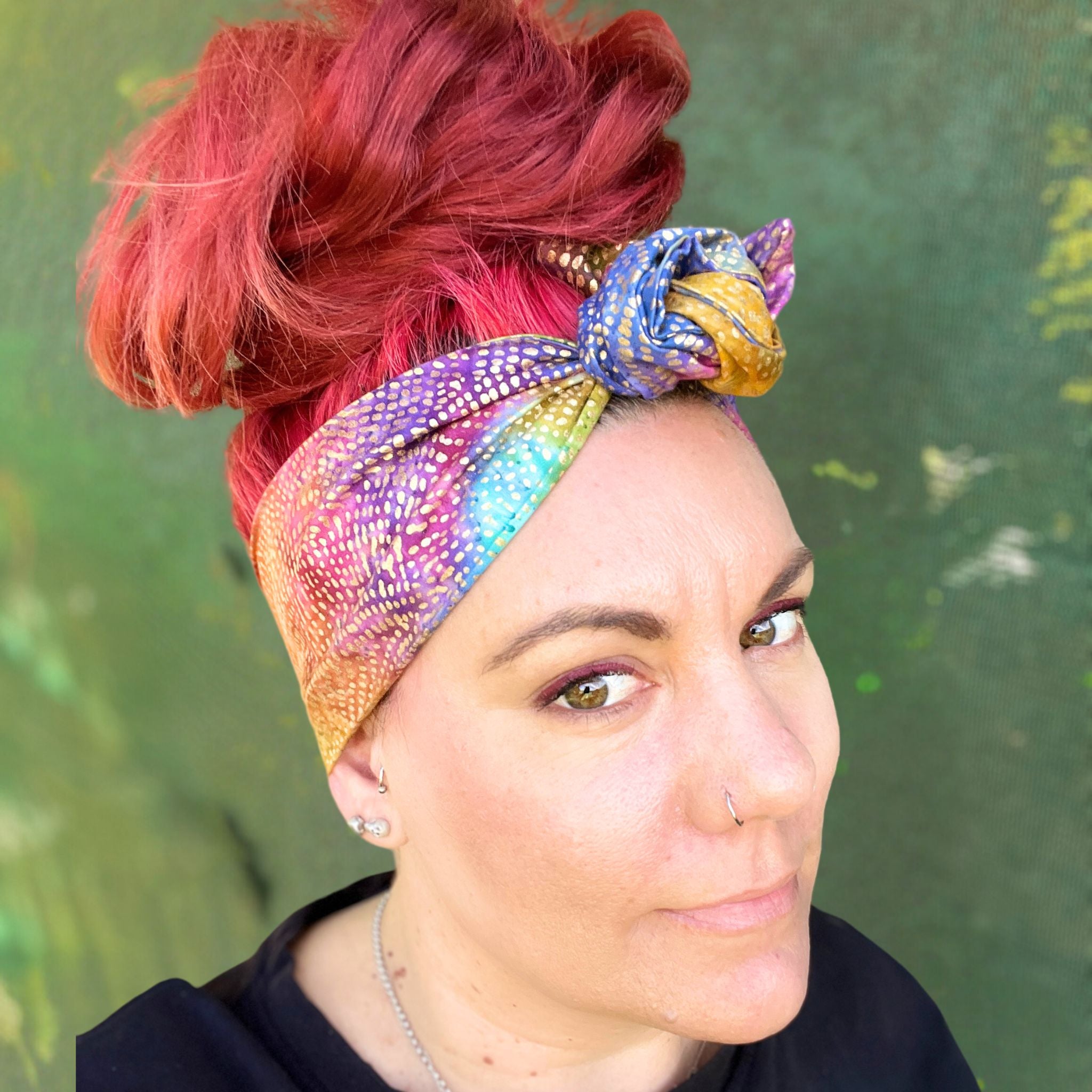 wire headwrap featuring a radiant metallic sheen with a gradient of colors, resembling a rainbow effect. The headwrap is artfully tied in a knot at the side of her head, lending a touch of whimsy to the overall look. 