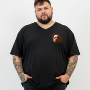 A man with a beard and tattooed arms wearing a black v-neck t-shirt with a small skull and red roses graphic on the left chest, standing against a light gray background.