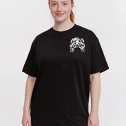 A woman wearing a black oversized short-sleeve t-shirt with a metallic silver and black graphic print on the left chest, set against a plain light gray background.