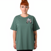 A woman with red hair in a bun wearing a sage green t-shirt with a black and white geometric patterned graphic on the left chest, set against a plain white background.