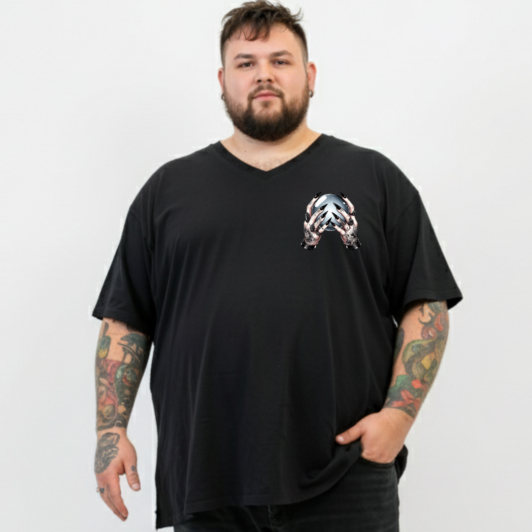 A man with a beard and tattooed arms wearing an oversized black V-neck t-shirt with a small metallic graphic on the left chest, standing against a light gray background.