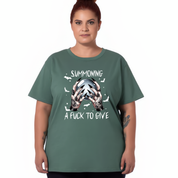 A woman modeling a sage green t-shirt with a graphic on the front showing hands holding a glowing orb with the text 'SUMMONING A FUCK TO GIVE' and small white bats.