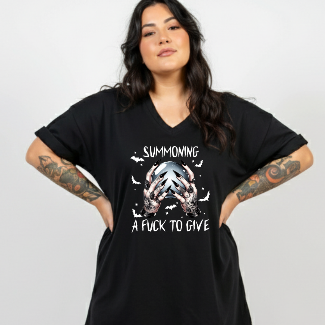 A woman with long dark hair and tattooed arms models a black oversized V-neck t-shirt with a white graphic print that reads 'SUMMONING A FUCK TO GIVE' with a skull and bat design.
