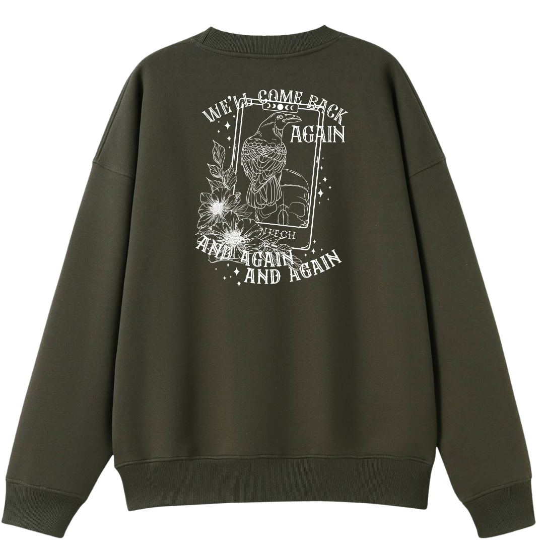 An army green oversized crew neck shown from the back, with the We'll Come Back Again graphic printed large across the back, a crow on a tarot card framed by flowers.